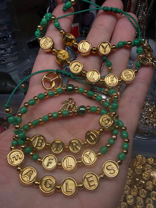 Green beaded bracelets with gold letter beads spelling Fly, Eagles, and Birds, held in a hand