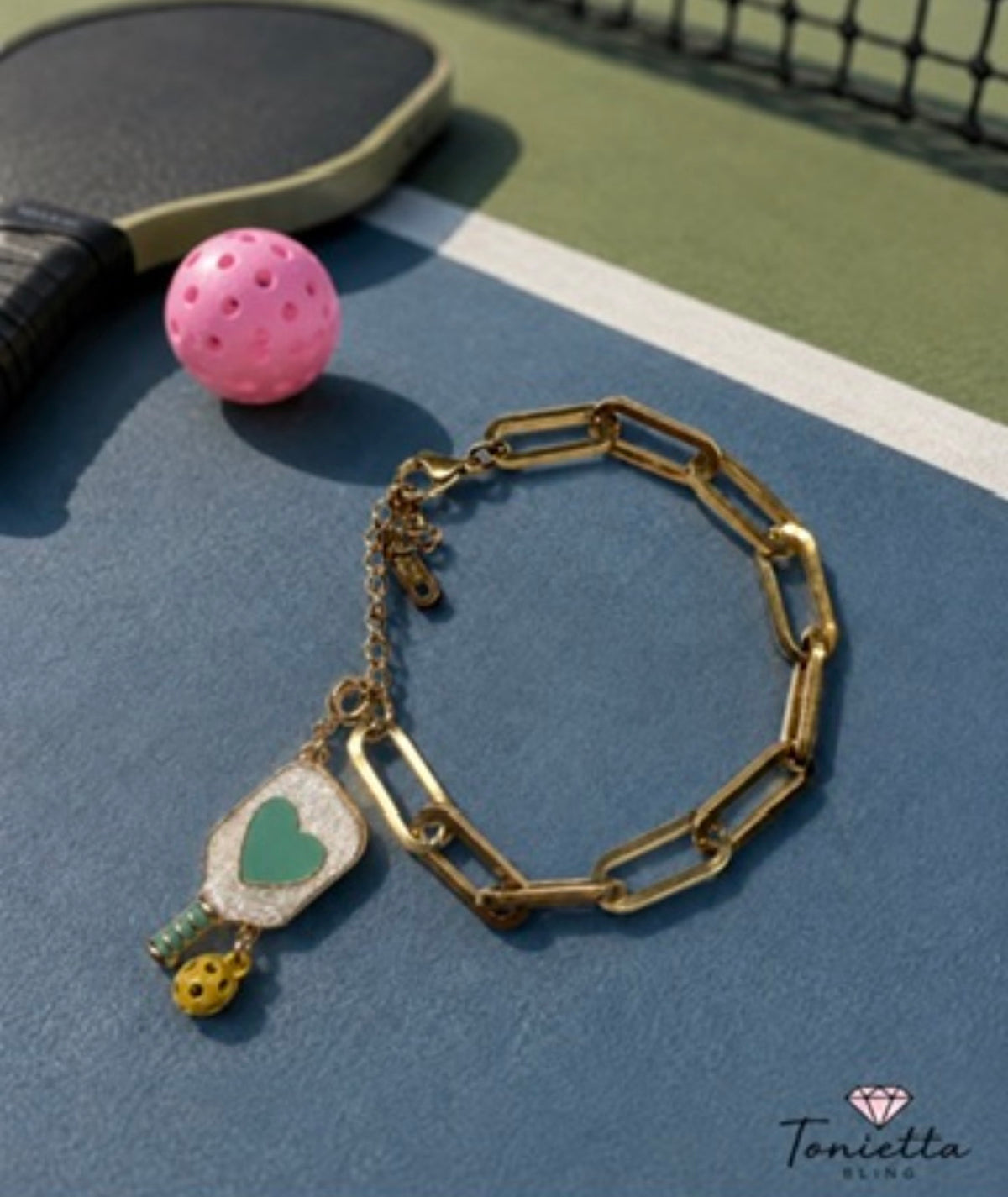 Gold paperclip chain bracelet with a glitter pickleball paddle charm with a green heart and a tiny gold pickleball, on a pickleball court